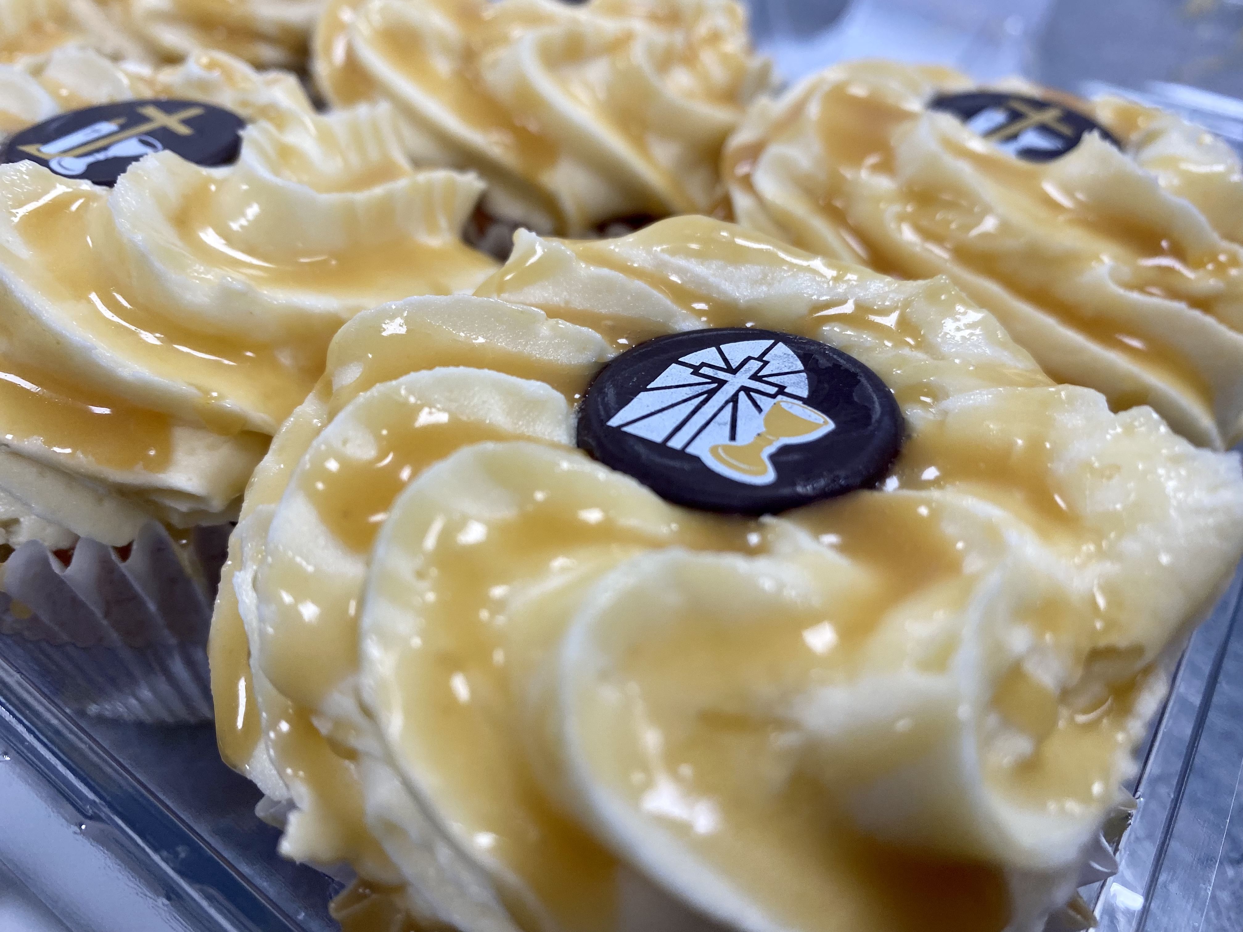 A close-up of several Salted Caramel Cupcakes with swirled salted caramel frosting. Each cupcake is topped with a decorative chocolate disk featuring a cross and a small golden footprint. The treats are arranged together in a clear plastic container.